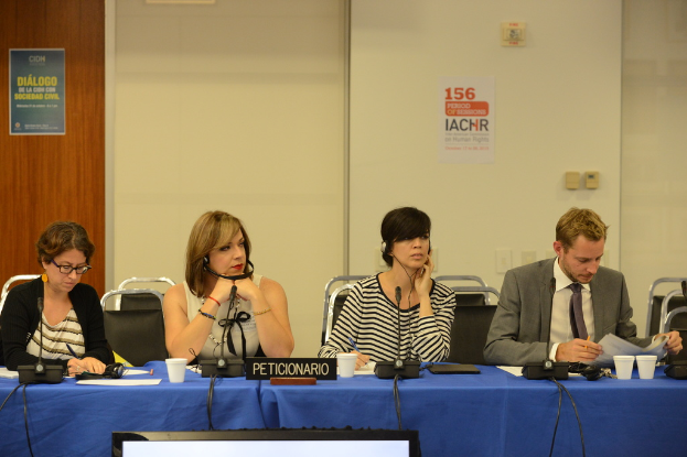 Ein Besprechungsraum mit einem blauen Tisch, vier sitzenden Personen, einer Tür, einer Wand und einem Plakat an der Wand.