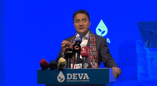 Ein Mann in einem Mantel und einer Mütze steht an einem Podium mit Mikrofonen und spricht zu einem Publikum. Das Podium hat ein Schild mit der Aufschrift "deva-Geschäftsführer sagt, es sei keine gute Idee für die Zukunft". Im Hintergrund gibt es ein Objekt und einen Bildschirm.