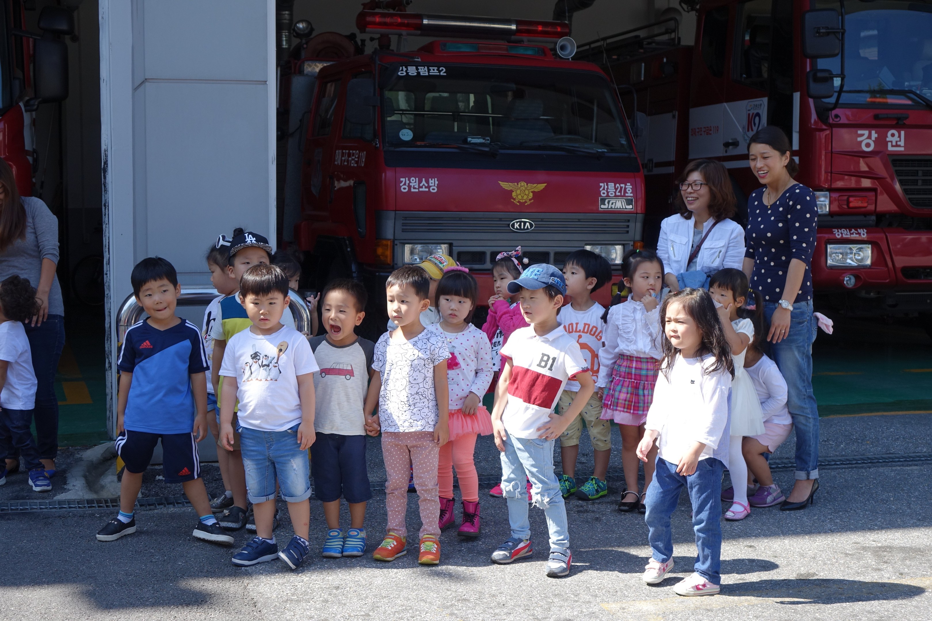Eine Gruppe von Kindern steht vor einem Feuerwehrauto auf einem Feuerwache-Hof, einige tragen Mützen, mit zusätzlichen Feuerwehrautos im Hintergrund.