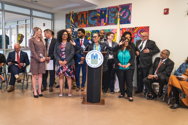 Gruppe von Menschen, die vor einem Rednerpult mit einem Mikrofon und einer Tafel mit der Aufschrift "Sancho City Councilman Joins City Council" stehen, mit sitzenden und stehenden Zuschauern, Flaggen, Feuerlöscher und Deckenleuchten im Hintergrund.