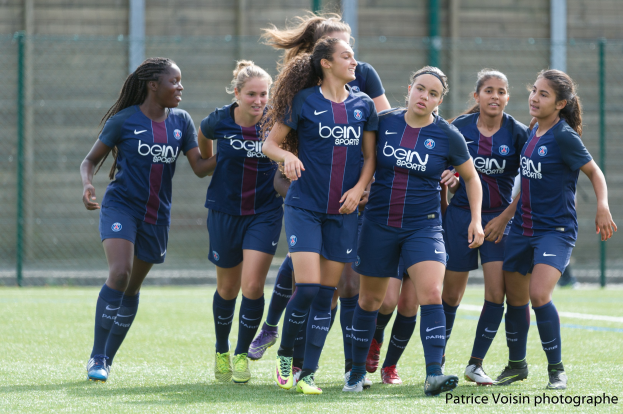 Gruppe junger Frauen beim Fußballspielen auf einem Rasenfeld mit Maschendrahtzaun und einer Wand im Hintergrund, beschriftet mit "Paris Saint-Germain Frauenfußball" in der rechten unteren Ecke.