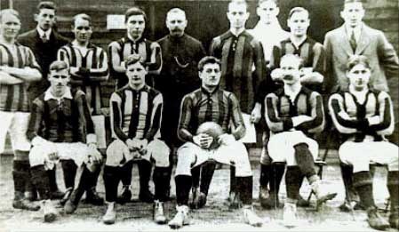 Schwarzes und weißes Foto einer Fussballmannschaft, einige stehen und einige sitzen, vor einer Wand mit dem Text "Ballymena Football Club" unten.