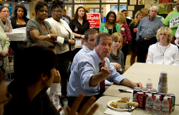 Eine Treffenszene mit einem Tisch im Vordergrund, der Coke-Dosen, Gläser, Wasserflaschen, einen Teller und Lebensmittel hält, einige Menschen in der Mitte sitzen, Glastüren im Hintergrund, ein Anschlagbrett oben rechts und ein Schreibtisch mit einigen Kisten links.