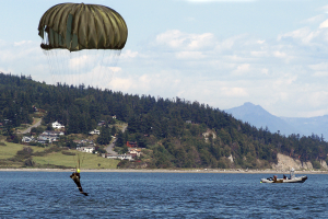 Eine Person gleitet mit einem Gleitschirm über einen See, mit einem Boot auf der rechten Seite, Bäumen, Gebäuden, Hügeln und einem klaren blauen Himmel im Hintergrund.
