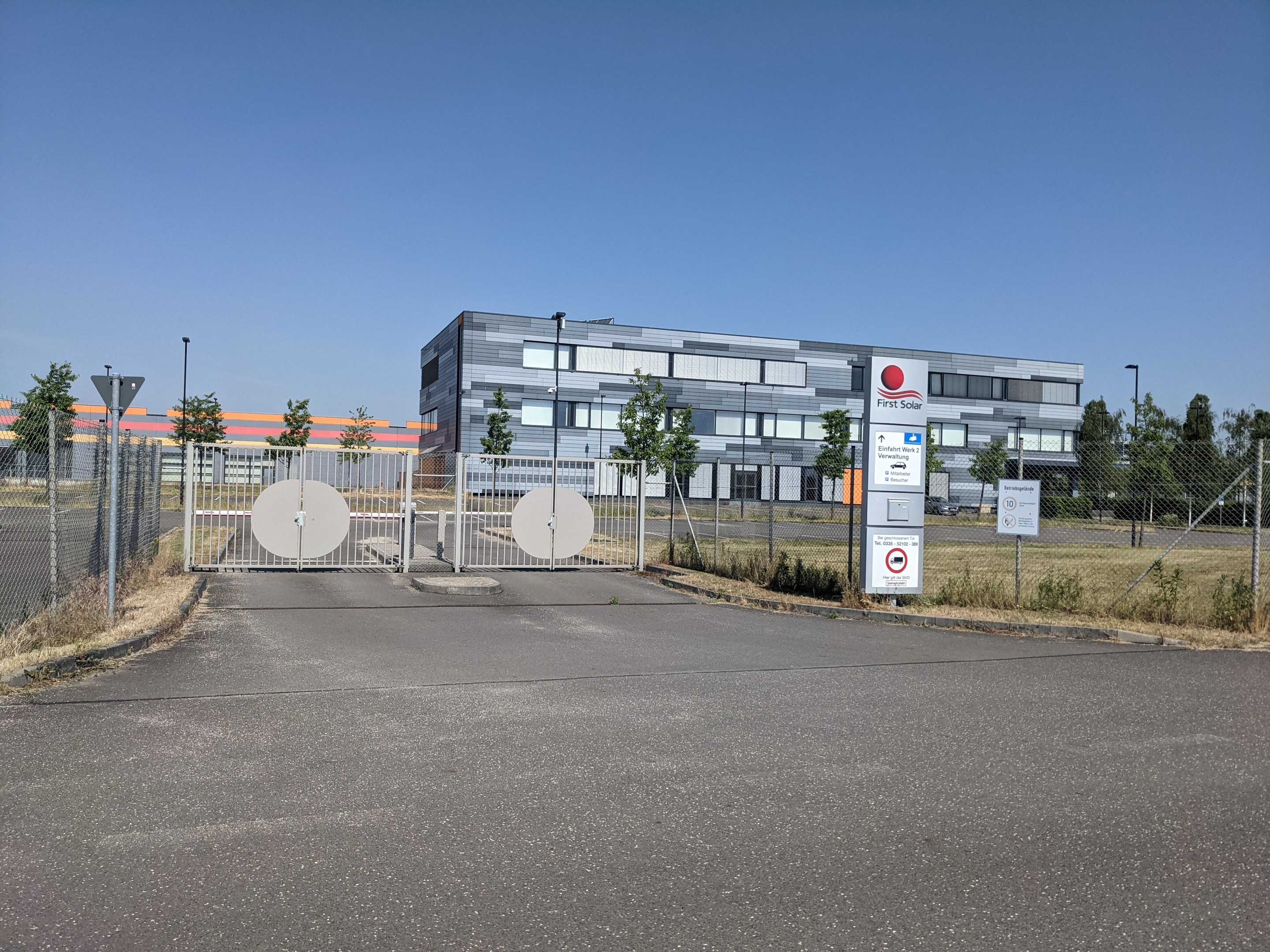 Großes industrielles Gebäude mit der Bezeichnung "First Solar" und vielen Fenstern, umgeben von einem Metallzaun, Bäumen, Pflanzen, Gras, Schildern, Straßenlaternen und einem Weg unter einem bewölkten Himmel.
