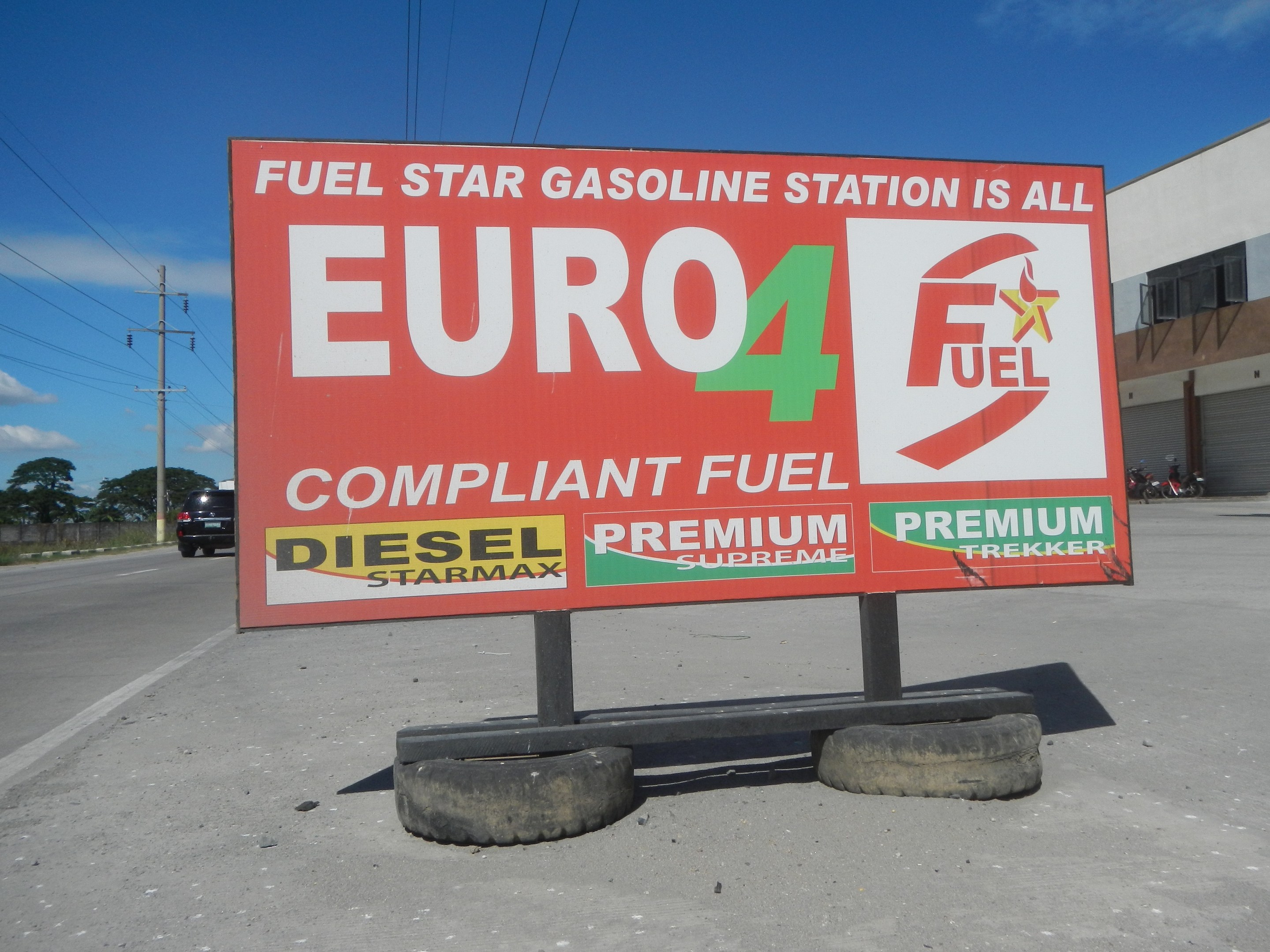 Tankstelle mit "Euro4 Fuel"-Schild im Vordergrund, Fahrzeuge auf der Straße, Strommäste mit Drähten, Bäume, Gebäude auf der rechten Seite und Himmel überall.