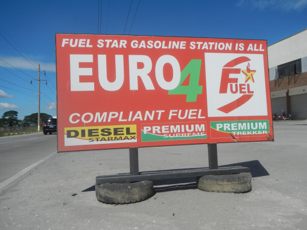 Tankstelle mit "Euro4 Fuel"-Schild im Vordergrund, Fahrzeuge auf der Straße, Strommäste mit Drähten, Bäume, Gebäude auf der rechten Seite und Himmel überall.