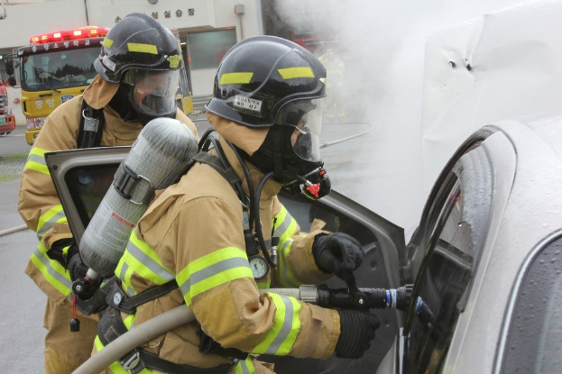 Feuerwehrleute in Schutzausrüstung verwenden einen Schlauch, um ein brennendes Auto zu löschen, mit Rauch und Fahrzeugen und einem Gebäude im Hintergrund.