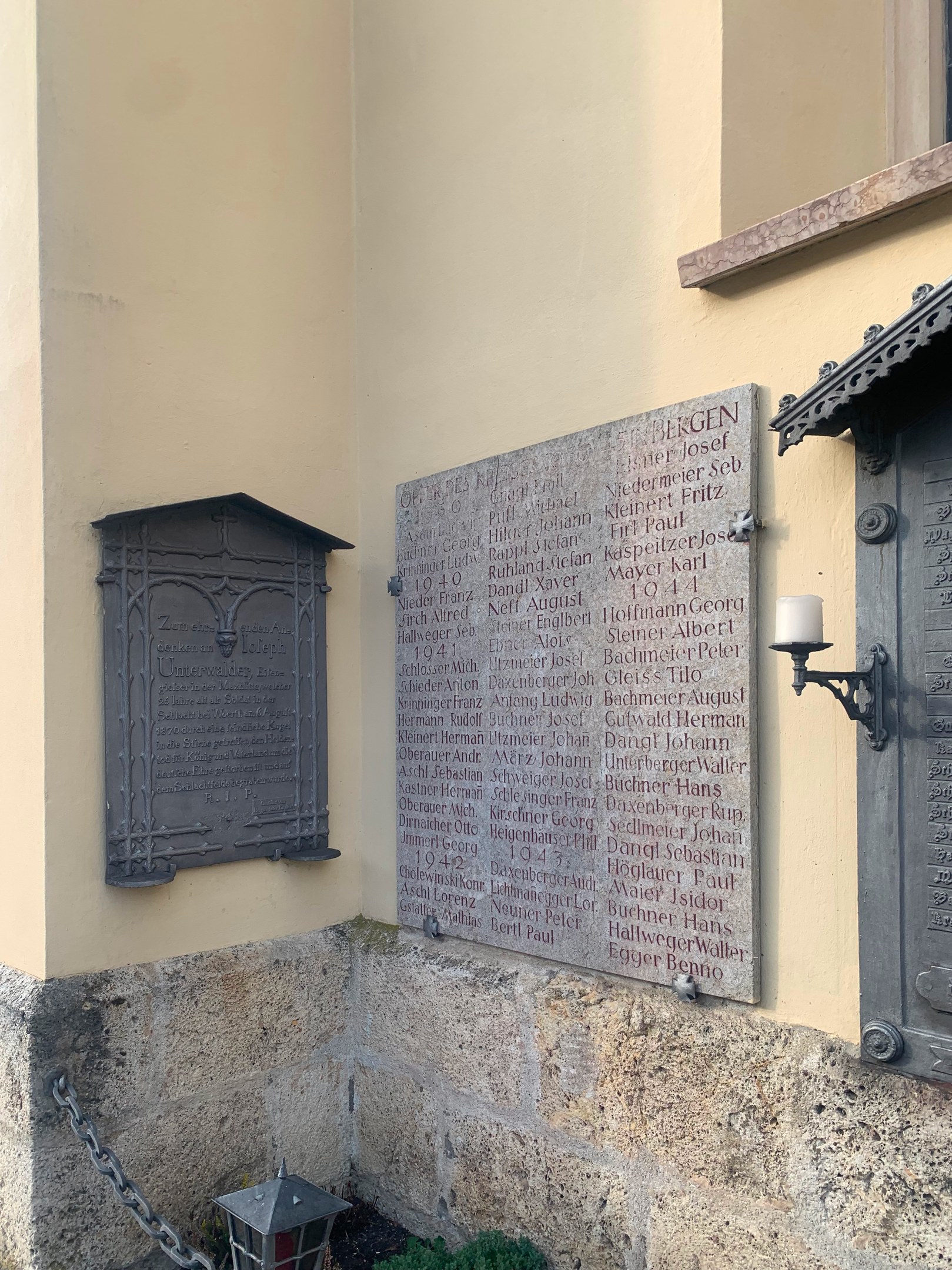 Ein Gebäude mit einer Gedenktafel, einem Fenster, einer Tür, einer Leuchte, einer Kette und Topfpflanzen.