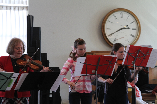Drei Frauen auf der Bühne stehend und Geigen spielend, mit Notenpulten und Noten davor, während das Publikum sitzt und zuhört; eine Uhr hängt an der weißen Wand im Hintergrund, und ein weißer Vorhang ist links zu sehen.