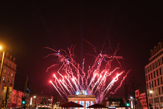 Eine belebte Stadtstraße in Berlin an Silvester, voller Menschen, Fahrzeuge und erhellt von Feuerwerk und Gebäudelichtern, die eine festliche Stimmung schaffen.