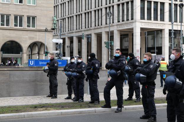 Eine Gruppe von Polizisten in schwarzen Uniformen und Masken steht vor einem Gebäude mit Glasfenstern und Säulen in Berlin, Deutschland, einige halten Helme, mit Laternen, Verkehrszeichen, Schildern mit Text, einer Statue, anderen Menschen und grasbewachsenem Boden im Hintergrund.
