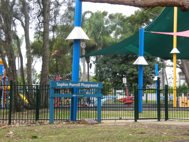 Ein Spielplatz mit grüner Überdachung und Metallzaun, der Kinder beim Spielen auf Geräten zeigt, ein Namensschild, Pfähle, Bäume, Gebäude und einen bewölkten Himmel.