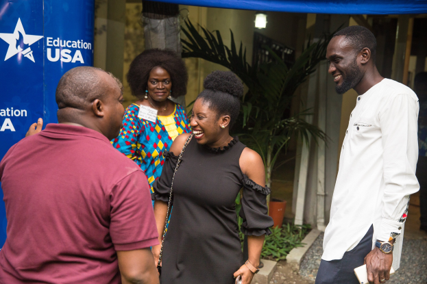 Gruppe von Menschen, die vor einem blauen Zelt mit einer Fahne, Topfpflanzen, einer Säule, Wand und einer Decke mit Leuchten sprechen.