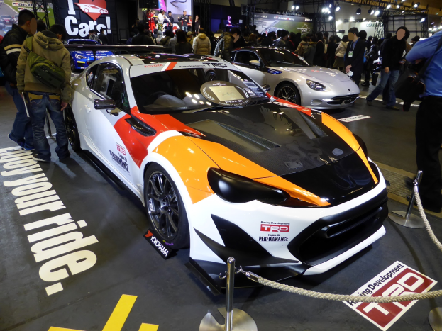 Eine Gruppe von Menschen um ein Rennfahrzeug auf der Tokyo Motor Show herumsteht, umgeben von Absperrpollern und Seilen, sowie Informationsschildern im Hintergrund unter beleuchteter Decke.