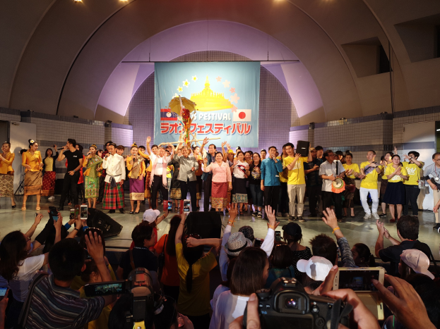 Eine Gruppe von Menschen auf einer Bühne mit Kameras, Handys und Musikinstrumenten während einer Festivaleröffnung, mit einem Banner, Wänden und Deckenlampen im Hintergrund.