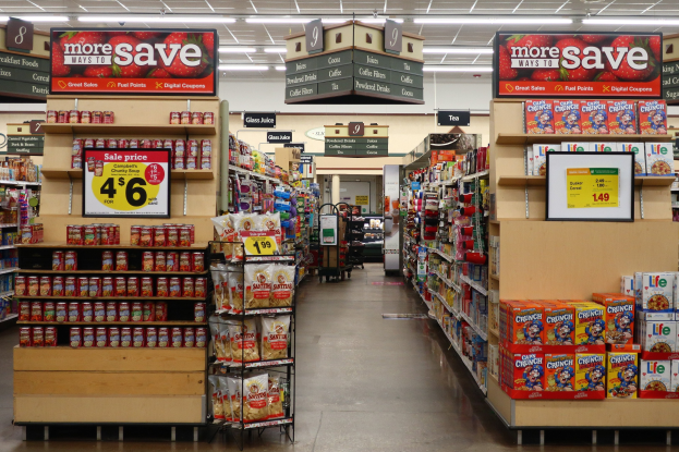 Ein Kiosk-Innenraum mit Regalen und Boden voller verschiedener Lebensmittel, Deckenbeleuchtung und einer Person im Hintergrund.