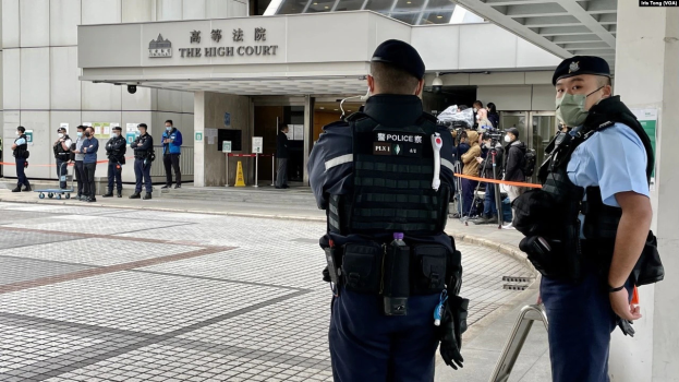 Zwei Polizisten in Uniform, Mützen und Masken stehen vor einem Gebäude am Hong Kong International Airport, umgeben von einer Gruppe von Menschen, einige halten Kameras, mit Stangen mit Bändern im Hintergrund.