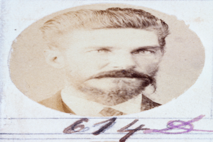 Ein altes Foto eines Mannes mit Bart und Schnurrbart, der direkt in die Kamera schaut und dessen Lippen leicht geöffnet sind, mit ordentlich gekämmtem Haar und einem gut gepflegten Bart.