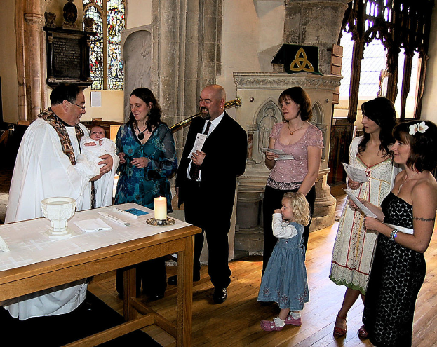 Ein Mann in einem weißen Kleid hält ein Baby, steht neben einer Frau in einem violetten Kleid, die das Baby ansieht, einem lachenden Mann im Anzug und einer Frau in einem pinken Hemd, die ein anderes Baby hält, mit zwei lachenden Frauen rechts und einer Wand, einem Fenster, einem Anschlagbrett und einem Schild im Hintergrund.