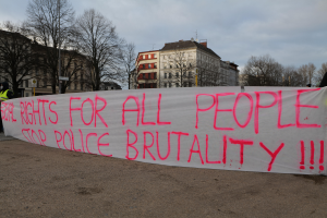 Eine Gruppe von Menschen, die auf dem Boden stehen und ein Banner halten, auf dem 'Rechte für alle Menschen Stoppt Polizeigewalt' steht, mit einem Straßenschild, einem Schild, Bäumen, Gebäuden mit Fenstern und einem bewölkten Himmel im Hintergrund.