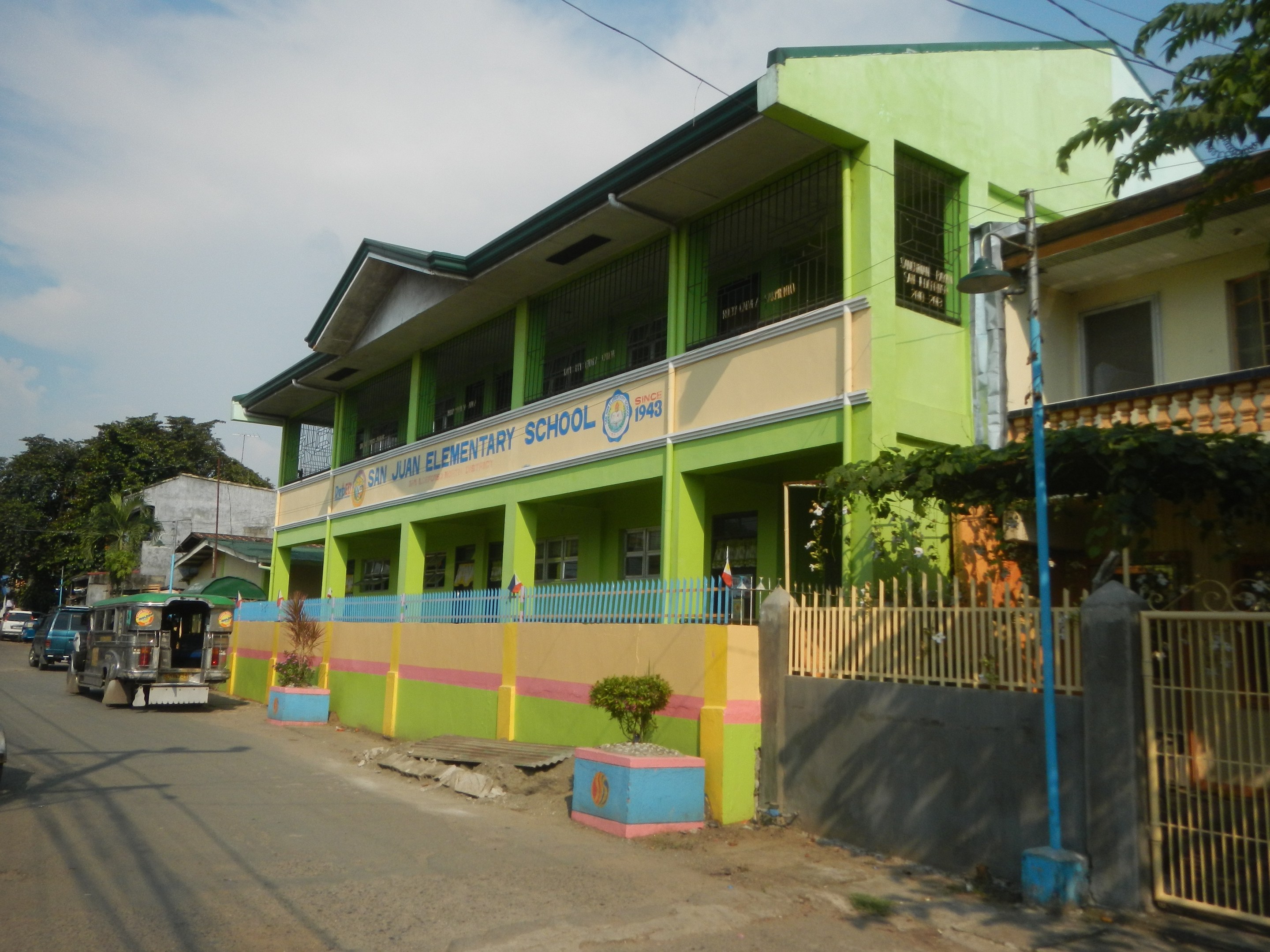 Schulgebäude mit grüner und gelber Fassade auf einer Straße mit Fahrzeugen, Blumentöpfen, einem Zaun, einem Tor, einem Straßenpfahl, Bäumen, Drähten und einem bewölkten Himmel.