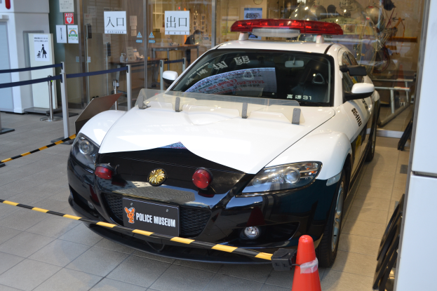 Ein Polizeiwagen ist vor einem Gebäude mit einem Verkehrskegel auf dem Boden, Absperrpollern mit Bändern, Hinweisschildern, einer Wand, einer Tür und einem Dach mit Deckenleuchten geparkt.