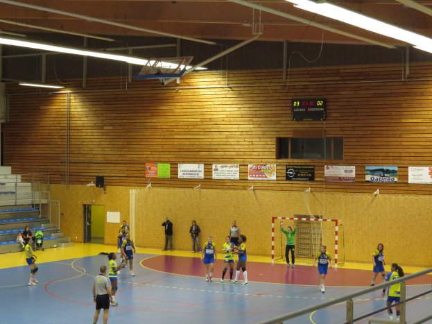 Eine Gruppe von Menschen, die Handball in einer Turnhalle mit einem Metallzaun, einem Tor, einer Tür, Texttafeln, einem Display, einem Lautsprecher, Metallstangen und einer Decke mit Deckenleuchten und Metallrahmen spielt.