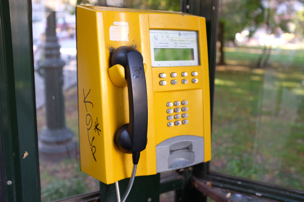 Ein gelbes Münztelefon auf einem Metallpfahl in der Vorderansicht, mit einem unscharfen Blick auf einen Pfahl, Bäume und Gras durch ein Glasfenster im Hintergrund.