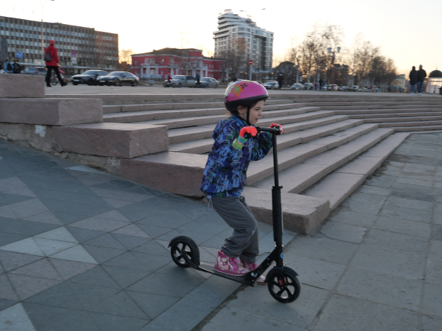 Ein mödchen in einem Helm fährt auf einem Scooter eine Treppe hinunter mit Fahrzeugen, Menschen, Laternen, Bäumen, Gebäuden und einem klaren blauen Himmel im Hintergrund.