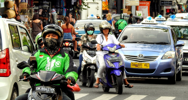 Eine Gruppe von Menschen auf Motorrädern fährt auf einer belebten Straße mit Autos und anderen Fahrzeugen, einige Fahrer tragen Helme, mit Gebäuden, Bäumen, Texttafeln und Geschäften im Hintergrund.