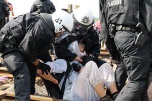 Eine Gruppe von Polizisten in schwarzer Einsatzkleidung verhaftet einen Mann in einem weißen Kleid auf einem Bahngleis, während andere Menschen in der Nähe stehen und der Himmel im Hintergrund zu sehen ist.