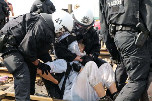 Eine Gruppe von Polizisten in schwarzer Einsatzkleidung verhaftet einen Mann in einem weißen Kleid auf einem Bahngleis, während andere Menschen in der Nähe stehen und der Himmel im Hintergrund zu sehen ist.