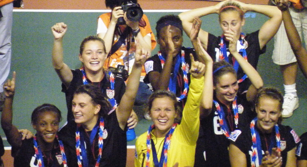Eine Gruppe von Frauenfußballspielerinnen auf einem Feld, die lächeln und Medaillen tragen, mit einem Mann im Hintergrund, der eine Kamera hält.