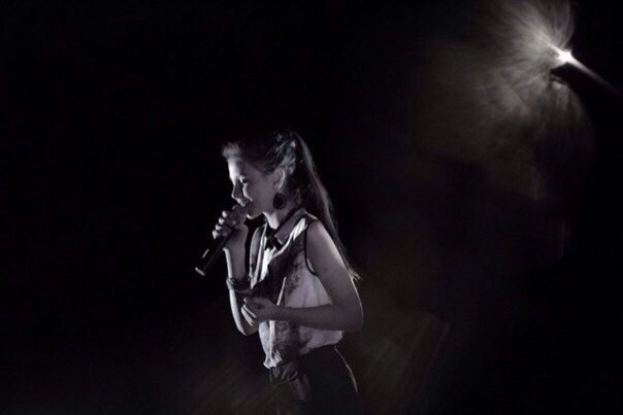 Eine Frau in einem schwarzen und weißen Outfit singt leidenschaftlich auf einer von Scheinwerfern beleuchteten Bühne, Mikrofon in der Hand, die Augen geschlossen.