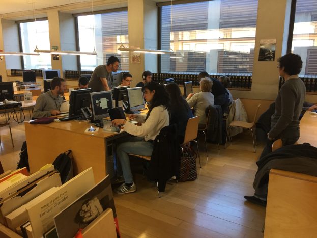 Menschen an Tischen in einer Bibliothek, die auf Computern mit Monitoren und Tastaturen arbeiten, umgeben von Bücherregalen und beleuchtet von Deckenlampen.