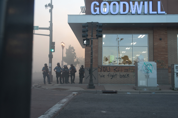 Eine Gruppe von Polizisten in Helmen und mit Waffen steht vor einem Goodwill-Laden mit Glaswänden, mit Laternenmasten, Verkehrsampeln, Bäumen und einem klaren Himmel im Hintergrund.