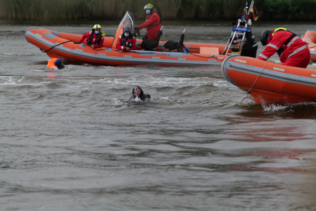 Eine Gruppe von Menschen in einem aufblasbaren Boot auf einem Fluss, mit zwei Personen im Wasser im Vordergrund und Vegetation im Hintergrund, alle mit Schwimmwesten und Helmen während einer Rettungsaktion.