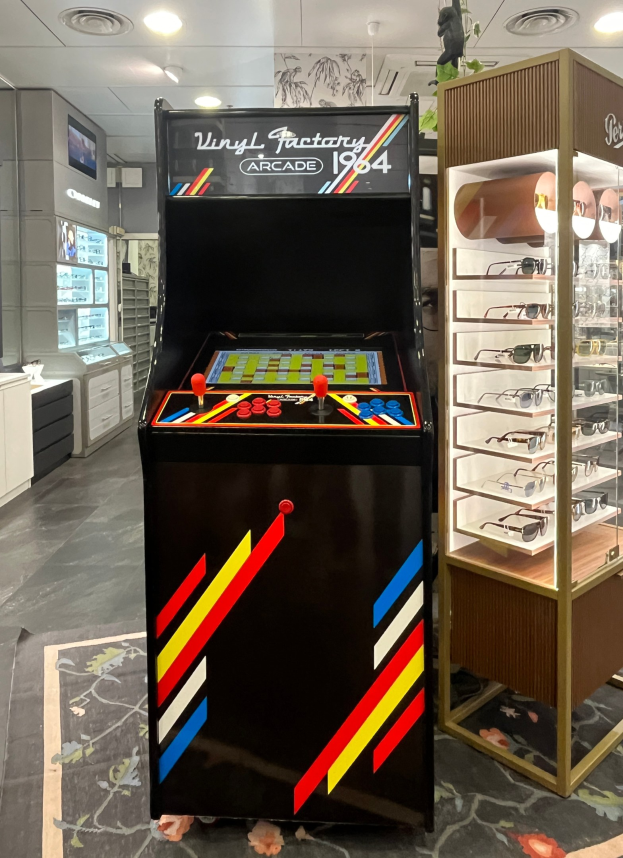Ein Retro-Laden mit einer alten Arcade-Maschine in der Mitte, Regalen, einem Fernseher und einer Brillenablage auf der rechten Seite unter Deckenlampen.
