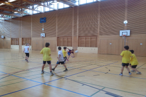 Kinder spielen mit einem Ball vor einer hölzernen Wand mit Basketballkörben, Holzstangen und Fenstern oben.