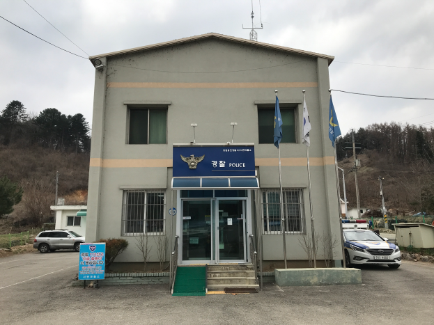Ein mehrstöckiges Gebäude mit Glasfenstern und -türen, Treppen mit Geländern, die zum Eingang führen, Fahnen an Masten, geparkte Fahrzeuge, ein Schild, Bäume im Hintergrund, Strommasten mit Drähten und ein bewölkter Himmel, identifiziert als das Polizeihauptquartier Seoul.