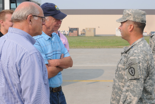 Eine Gruppe von Menschen, einige in Uniformen, die zusammen mit Kisten, Gras und anderen Gegenständen im Hintergrund und einer Straße mit markierten Linien im Vordergrund stehen.