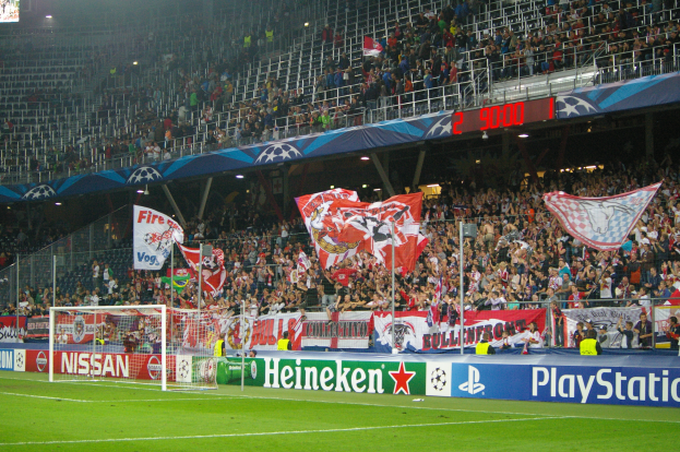 Ein Fussballspiel in einem hell erleuchteten Stadion mit einer grossen Zuschauermenge, grünem Rasen, zentralem Torpfosten mit Bannern und einem Bildschirm in der oberen linken Ecke.