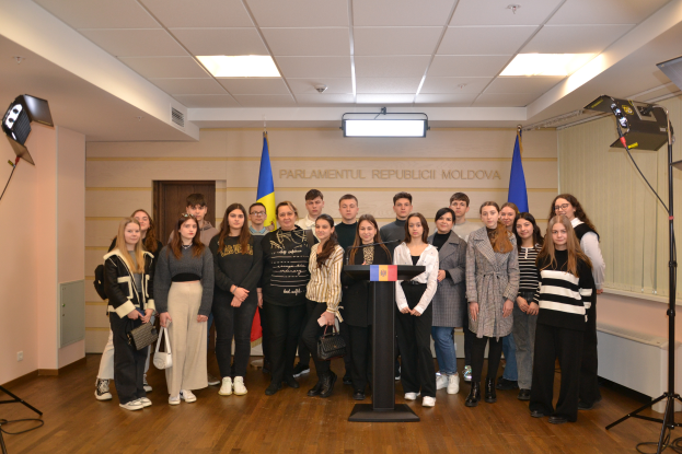 Eine Gruppe von Menschen steht vor einem Podium mit Mikrofon, umgeben von Fahnen, einer Wand mit dem Text "Moldova Jugendkongress 2018", einer Tür, einem Dach mit Deckenleuchten und Ständern mit Leuchten.
