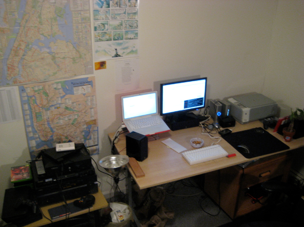 Ein Raum mit einem Tisch, auf dem ein Laptop, ein Lautsprecher, ein Monitor, eine Tastatur, ein Ball, eine Maus, ein Scanner und Papiere stehen, sowie ein weiterer Tisch mit elektronischen Geräten, Charts und Poster an der Wand dahinter.