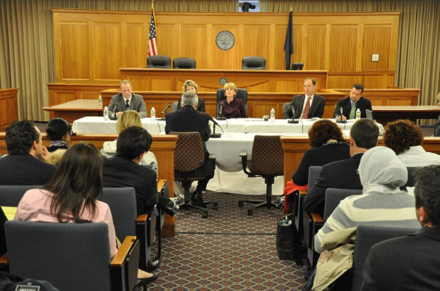 Ein Versammlungsraum mit einer Gruppe von Menschen, die auf Stühlen sitzen, zwei ausländische Flaggen, ein oranger Schreibtisch mit Wasserflaschen und ein Mikrofon.