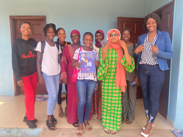 Eine Gruppe von Frauen steht vor einem Gebäude, lächelt und hält Bücher in den Händen, mit einem Tuch auf dem Boden rechts und Türen und einer Wand im Hintergrund, was auf eine Schule in Nigeria hindeutet.