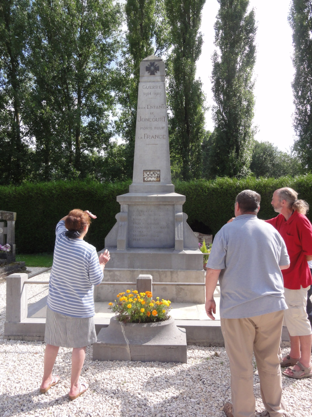 Eine Gruppe von Menschen, die um ein Weltkriegsdenkmal auf einem Friedhof stehen, mit einer Säule, die mit Text beschriftet ist, umgeben von Blumentöpfen, Bäumen und einem klaren blauen Himmel.