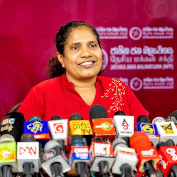 Eine Frau in einem roten Hemd sitzt an einem Tisch mit Mikrofonen, lächelt selbstbewusst, mit einem Banner dahinter, auf dem "Sri Lankas Minister für Gesundheit und soziale Dienste" steht.