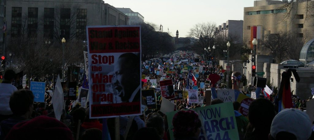 Menschen protestieren auf einer Straße und halten Schilder in der Hand, mit Gebäuden, Bäumen, Laternenmasten und dem Himmel im Hintergrund.